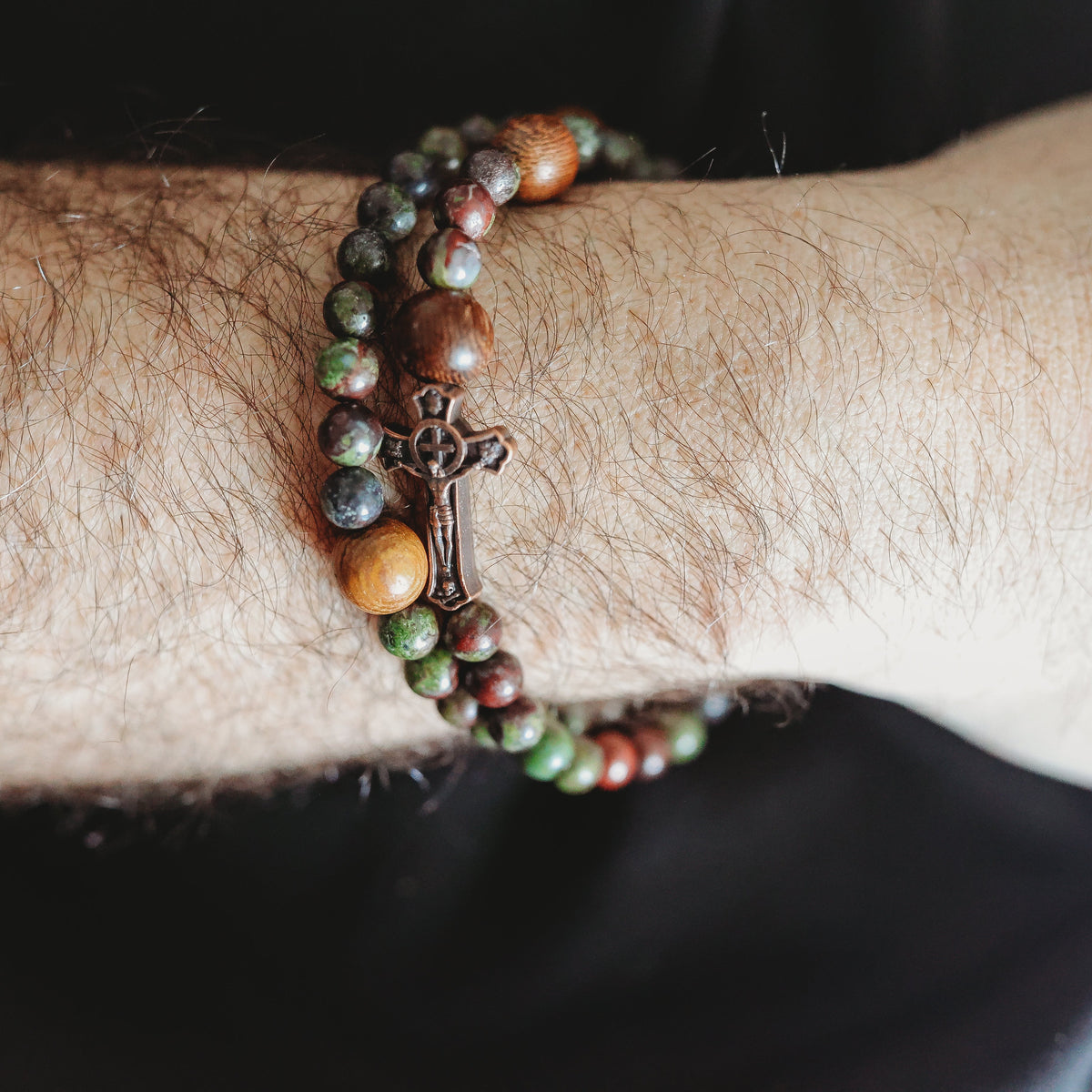 Close-up of a wrist wearing a beaded bracelet with a cross charm on a dark background